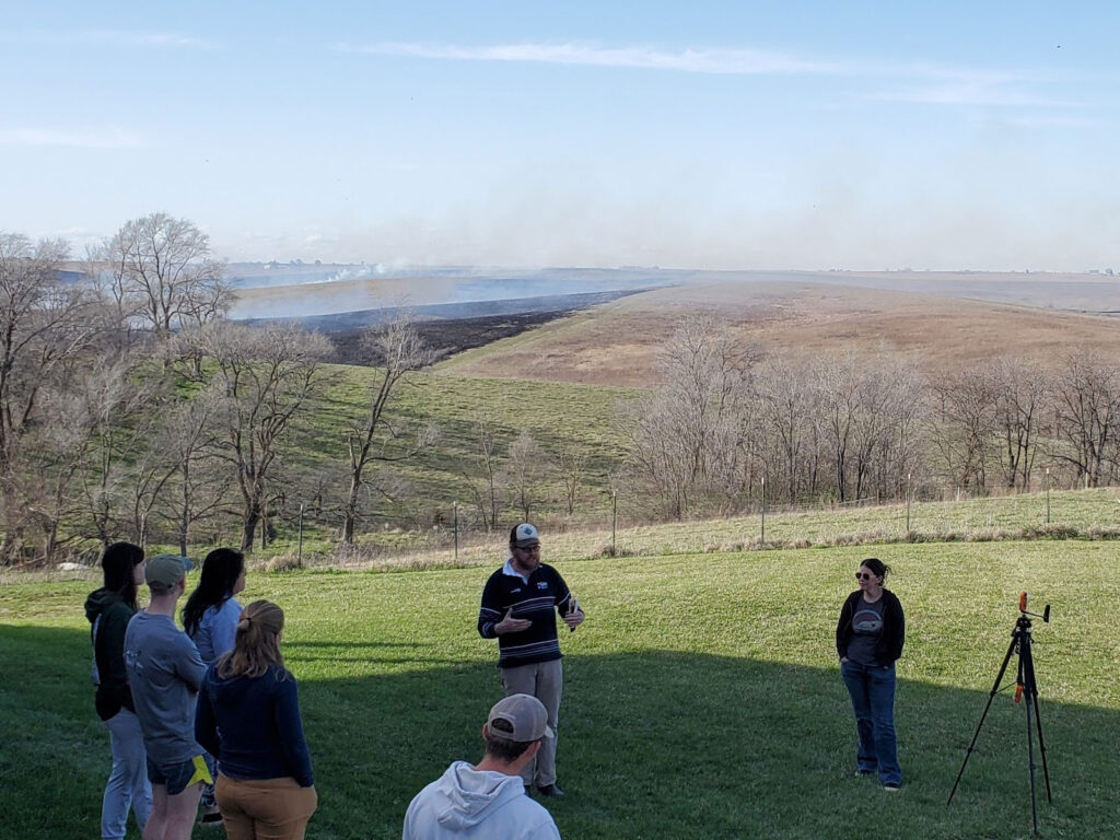 A speaker addressing a group of people on a grass covered hill with a controlled burn in the very distant background