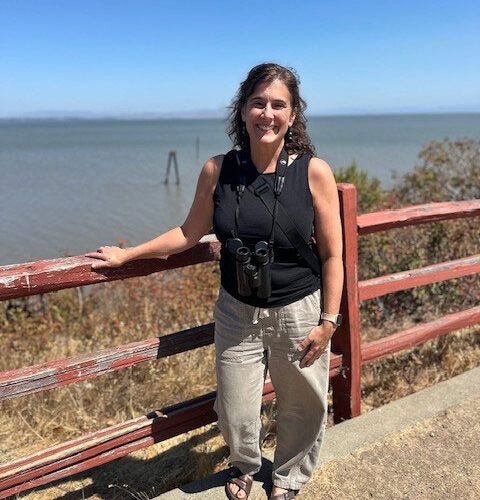 A woman leaning against a red wooden three railed fence with a body of water in the background