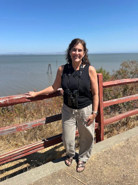 A woman leaning against a red wooden three railed fence with a body of water in the background